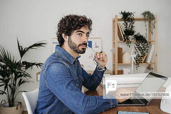 Contemplative engineer holding eyeglasses with laptop on desk in office