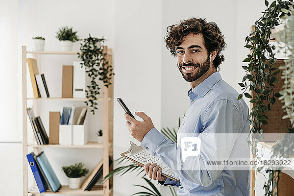 Happy businessman with smart phone holding blueprint in office