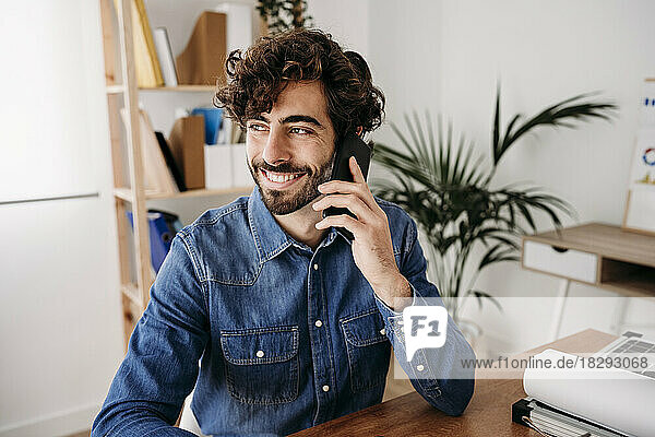 Glücklicher junger Ingenieur  der am Schreibtisch im Büro sitzt und mit dem Mobiltelefon spricht