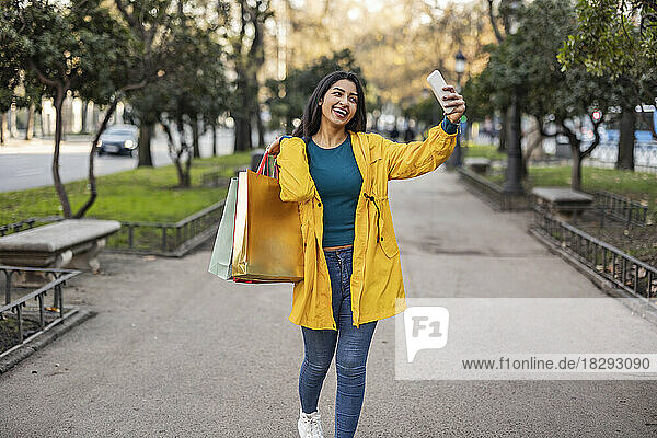 Happy woman with shopping bags taking selfie over mobile phone on footpath