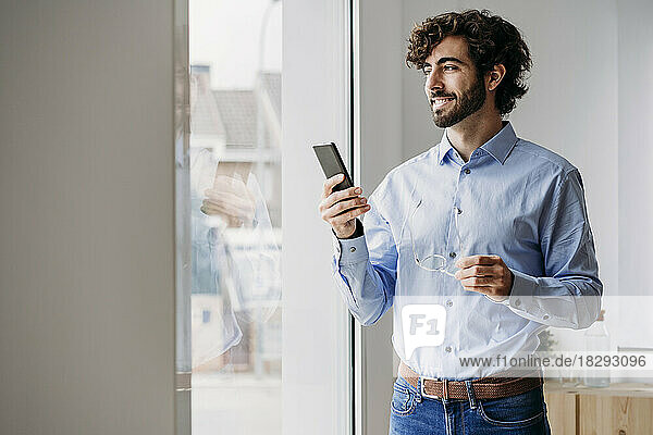 Happy businessman with smart phone looking through window in office