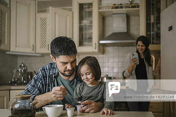 Father and son preparing tea with mother using smart phone in background