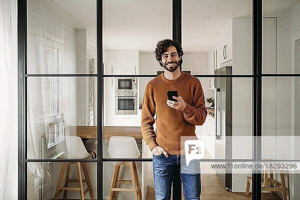 Happy man with smart phone leaning on glass door at home