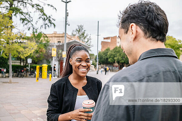 Happy young businesswoman holding disposable cup talking with colleague on footpath