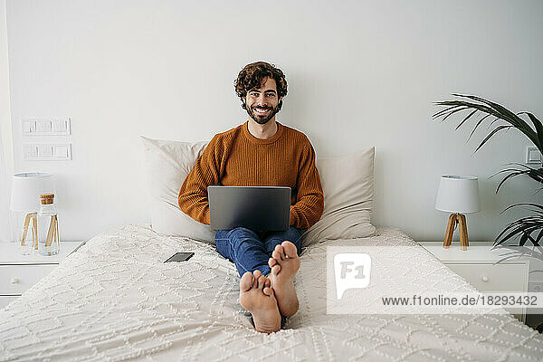 Happy young freelancer sitting on bed with laptop