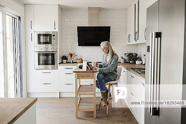 Reife Frau sitzt zu Hause in der Küche und benutzt einen Tablet-PC