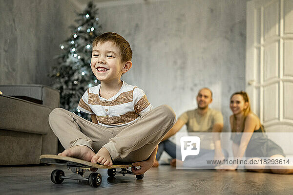 Glücklicher Vater und Mutter spielen mit ihrem Sohn  der zu Hause auf dem Skateboard sitzt
