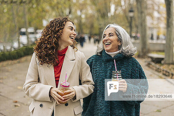 Happy young woman with grandmother holding glass of juice and walking on footpath