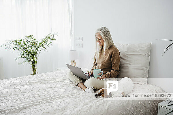 Reife Frau hält Kaffeetasse in der Hand und benutzt Laptop zu Hause