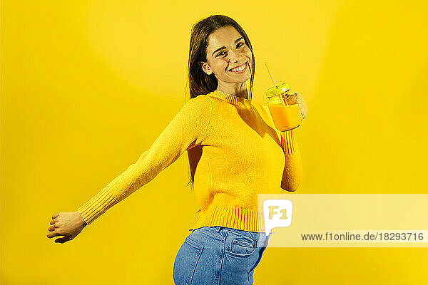 Glückliche junge Frau hält ein Glas Orangensaft vor gelbem Hintergrund