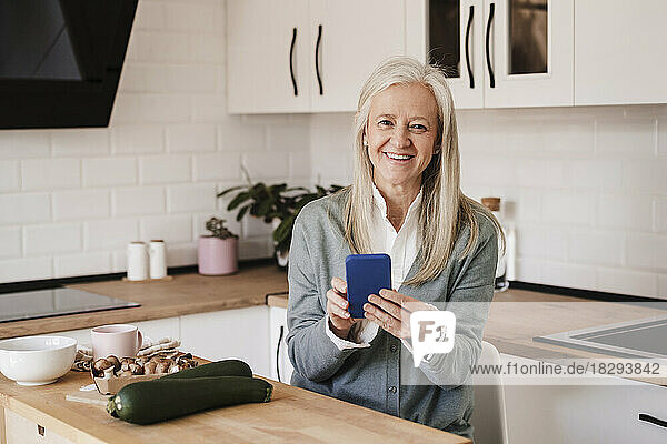 Glückliche reife Frau mit Smartphone sitzt zu Hause in der Küche