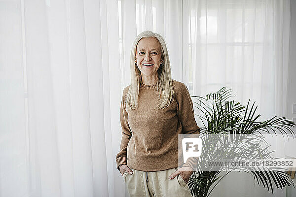 Happy mature woman with hands in pockets standing at home