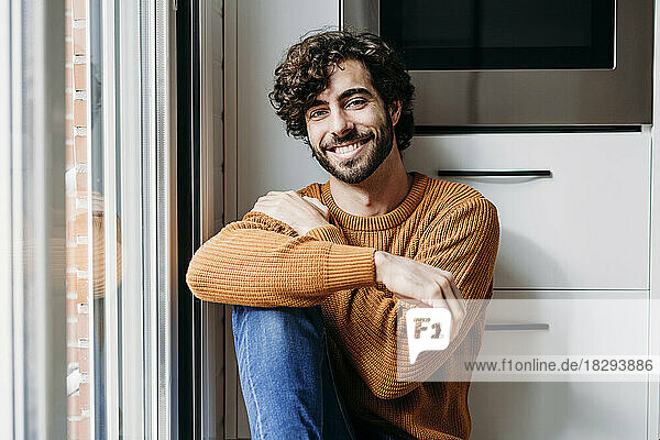 Lächelnder junger Mann  der zu Hause am Fenster in der Küche sitzt
