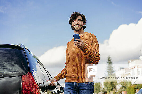 Glücklicher junger Mann mit Handy-Ladeauto an der Ladestation für Elektrofahrzeuge