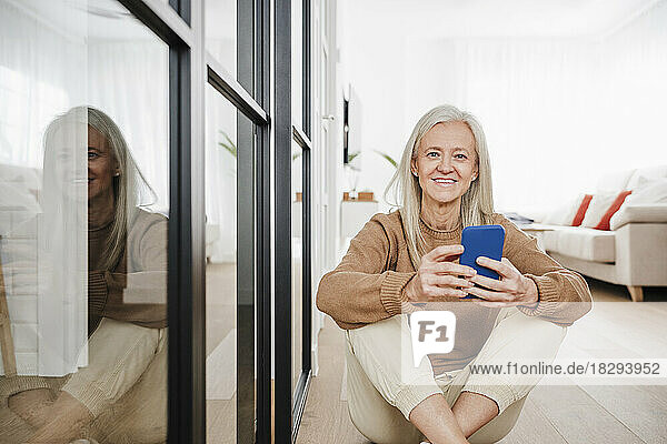 Glückliche Frau mit Smartphone sitzt zu Hause auf dem Boden im Wohnzimmer