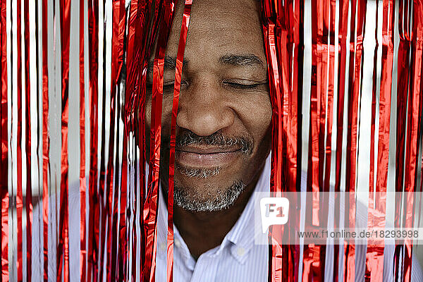 Lächelnder Mann mit geschlossenen Augen inmitten eines roten Lametta-Vorhangs