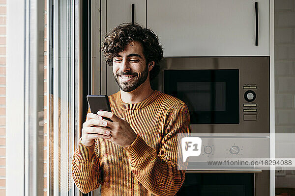 Happy young man using smart phone in kitchen at home