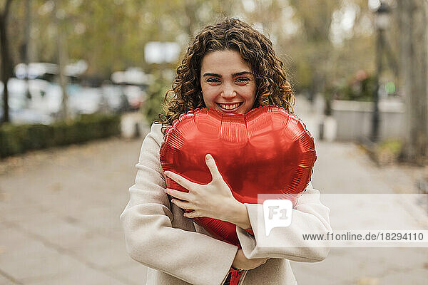 Happy young woman hugging red heart shaped balloon at footpath