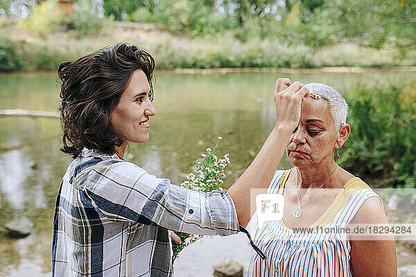 Smiling young woman putting flower on friend's head by lake in park