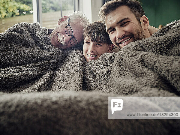 Happy grandfather  father and son snuggling under a blanket on couch in living room