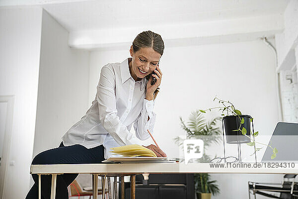 Glückliche reife Geschäftsfrau  die im Büro Notizen schreibt und über das Mobiltelefon spricht