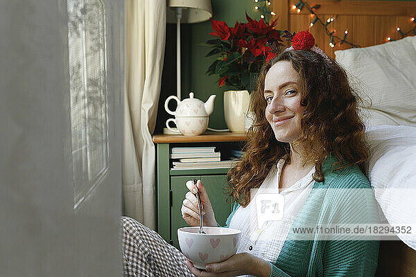 Smiling woman sitting with bowl by bed at home