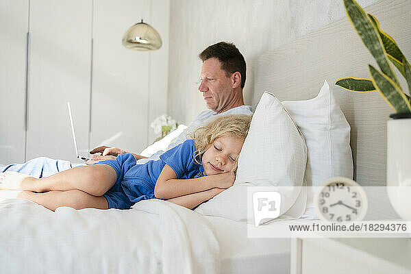 Daughter sleeping by father using laptop on bed in bedroom