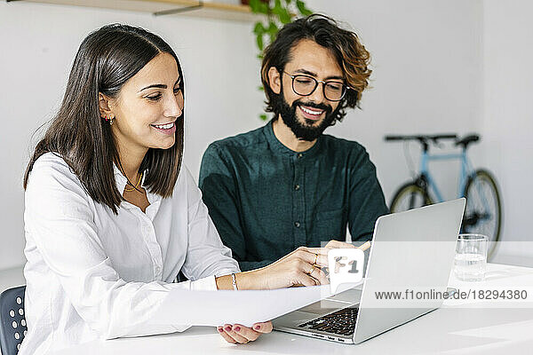 Happy businesswoman with document pointing at laptop by colleague in office