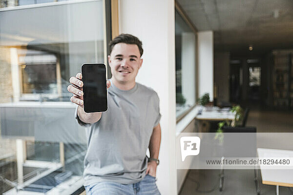 Smiling young businessman showing smart phone sitting on window sill in office