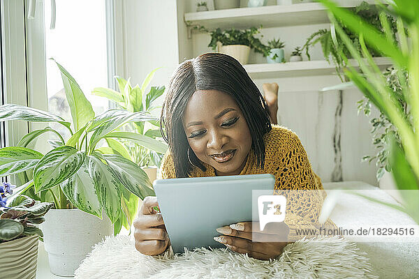 Smiling woman using tablet PC lying by window at home