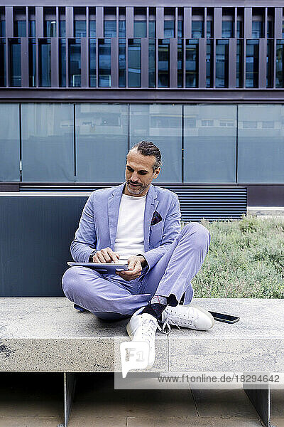 Reifer Geschäftsmann sitzt mit am Knöchel gekreuzten Beinen mit Tablet-PC auf einer Bank