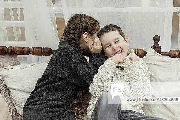 Sister whispering in brother's ear on sofa at home