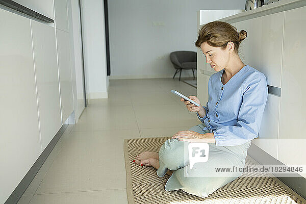 Woman using smart phone sitting by counter in kitchen