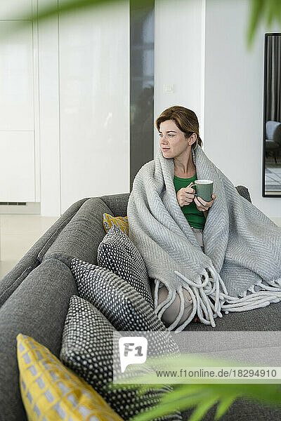 Contemplative woman wrapped in blanket sitting on sofa at home