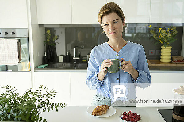 Woman holding coffee cup with croissant on counter in kitchen