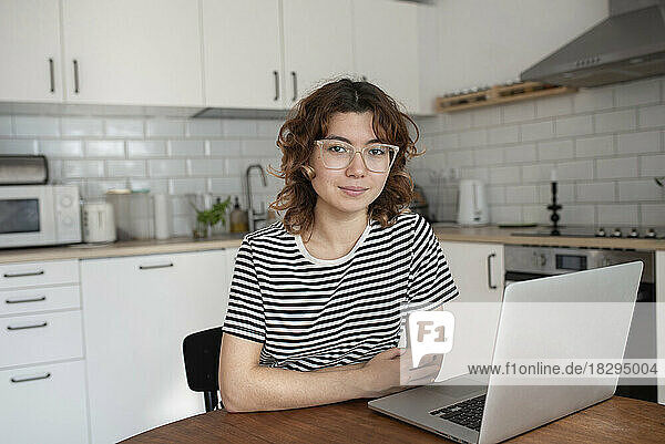 Young freelancer sitting with laptop at table in kitchen