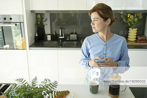 Thoughtful woman with smart phone standing by counter in kitchen