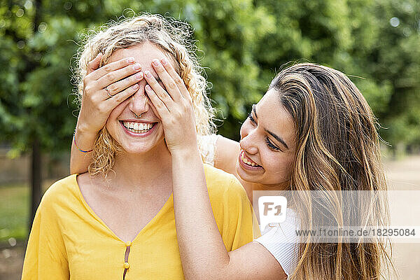 Smiling woman covering eyes of friend with hands in park
