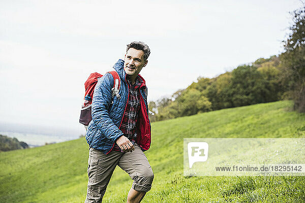 Smiling mature man with backpack walking on grass under sky