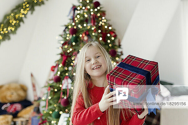 Glückliches blondes Mädchen hält Weihnachtsgeschenk vor dem Weihnachtsbaum
