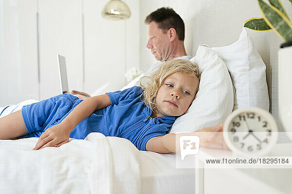 Girl holding alarm clock lying on bed by father in bedroom