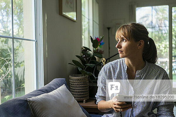 Frau mit Smartphone blickt zu Hause durch das Fenster