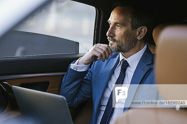Thoughtful businessman sitting with laptop in backseat of car