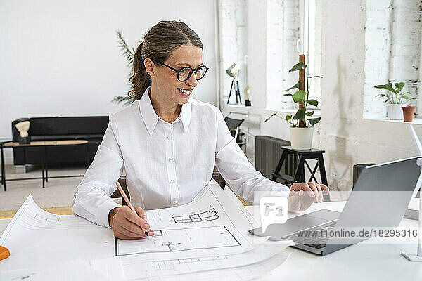 Happy businesswoman using laptop with blueprint in office