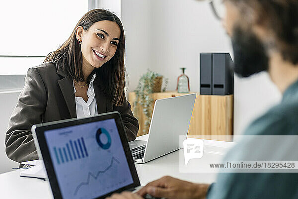 Smiling young businesswoman with laptop looking at colleague in office