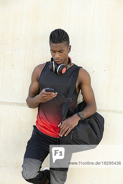 Young man holding gym bag using smart phone leaning on wall