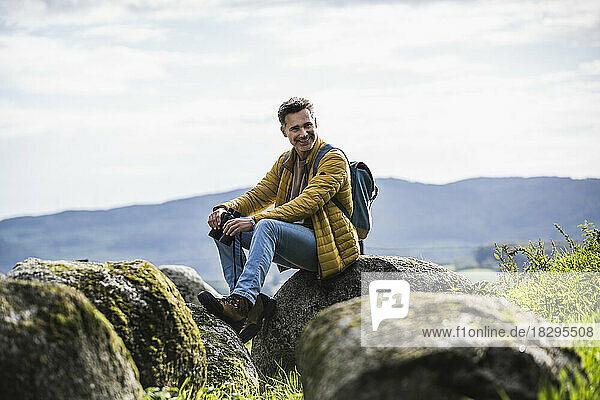 Happy mature man with backpack sitting on rock in front of sky