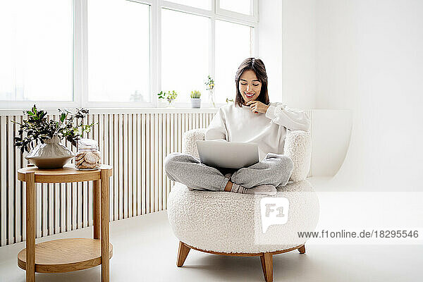 Lächelnde junge Frau  die zu Hause auf einem Stuhl sitzt und einen Laptop benutzt