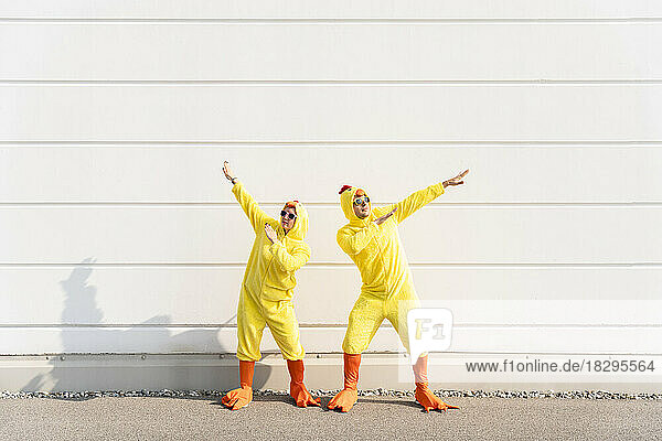 Verspielte Männer und Frauen in Hühnerkostümen gestikulieren und haben Spaß vor der Wand