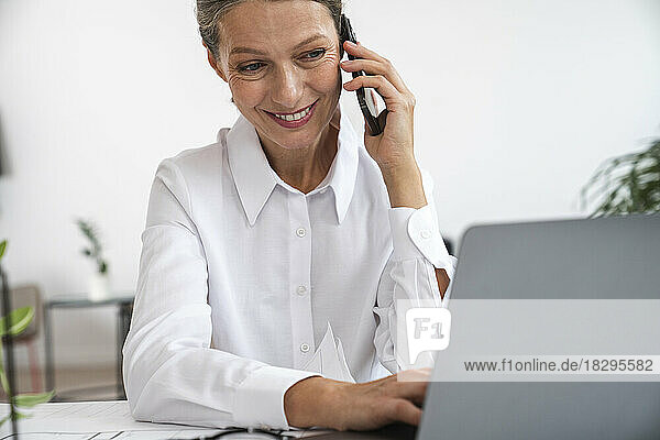 Happy mature businesswoman talking over mobile phone using laptop in office